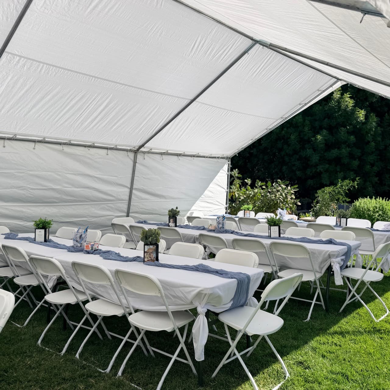 Long tables with white covers, dusty-blue runners, and small green centerpieces surrounded by white folding chairs beneath a white canopy on grass.